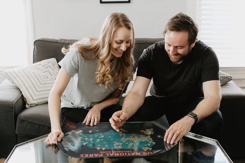 May include: A couple sitting on a sofa, looking at a map of the United States National Parks. The map is spread out on a glass coffee table. The map is labelled "US National Parks Explorer's Guide".