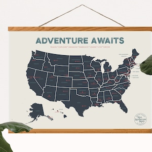 May include: A map of the United States with each state outlined in dark blue. The states are labeled with their names in white text. The map is titled "Adventure Awaits" with the tagline "Travel * Explore * Awaken * Embrace * Learn * Live * Grow". The map is printed on a cream-colored background and framed with a light brown wood frame.