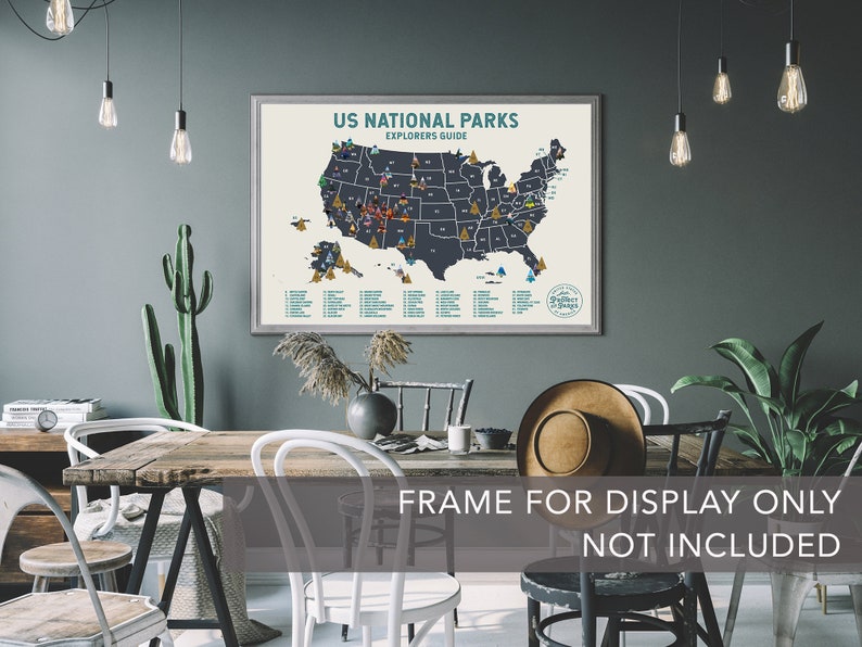 May include: A framed poster featuring a map of the United States, highlighting the locations of all the National Parks. The map is dark blue with white outlines and the text "US National Parks Explorers Guide" at the top. The poster is displayed on a grey wall in a room with a wooden table and chairs.