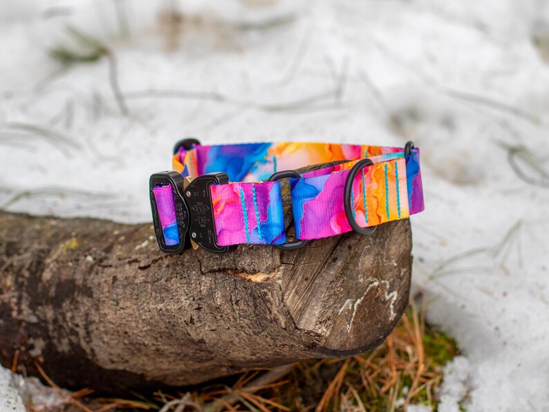 Puede incluir: Un collar para perros colorido con un dise&ntilde;o vibrante de acuarela en tonos de azul, rosa, naranja y morado. El collar tiene una hebilla y una anilla en D de metal negro. El collar est&aacute; sobre un trozo de madera, con un fondo nevado.