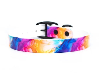 May include: A white dog collar with a colorful, abstract design. The collar features a black buckle and a vibrant pattern of pink, orange, blue, and yellow hues. The collar is designed for pets.