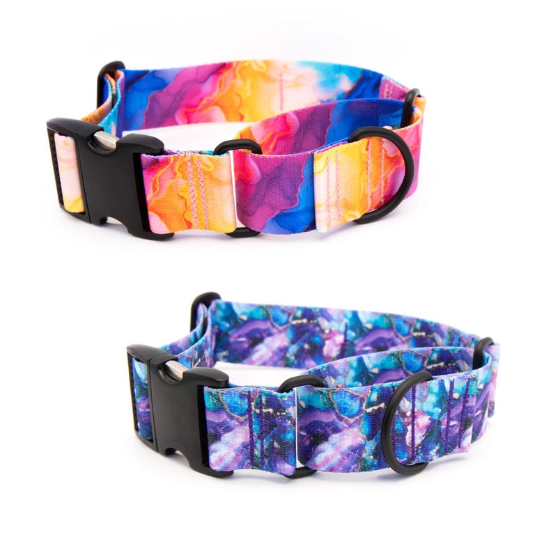 May include: Two pet collars featuring vibrant, abstract patterns. The top collar displays a blend of pink, orange, yellow, and blue hues. The bottom collar features a deep purple, blue, and teal design. Both include black plastic buckles and metal D-rings.