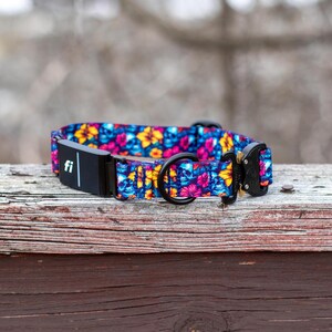 May include: A dog collar featuring a vibrant pattern of blue skulls and yellow and pink flowers. The collar includes a black electronic tracking module with the text fi, a black metal D-ring for a leash, and a black plastic side-release buckle.