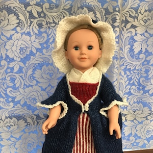 May include: A doll wearing a blue and white striped dress with a red and white striped skirt, a blue jacket, and a white bonnet.