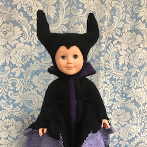Puede incluir: Una muñeca vestida como Maléfica de la película de Disney La Bella Durmiente. La muñeca lleva un vestido negro de punto con detalles morados y un sombrero a juego con cuernos.