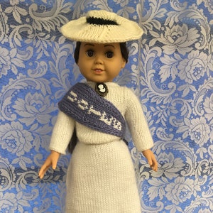 May include: A white knitted doll wearing a white hat with a black band, a white sweater, a purple and white scarf, and a white skirt. The doll is standing on a white stand.