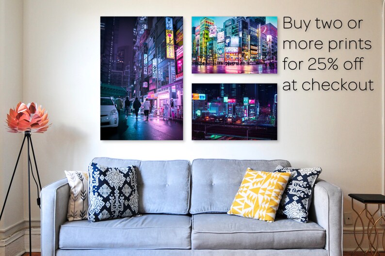 Pu&ograve; includere: Tre stampe incorniciate di paesaggi urbani notturni con luci al neon. Le stampe sono appese a una parete bianca sopra un divano grigio con cuscini. Il testo "Acquista due o pi&ugrave; stampe con il 25% di sconto alla cassa" &egrave; nell'angolo in alto a destra dell'immagine.