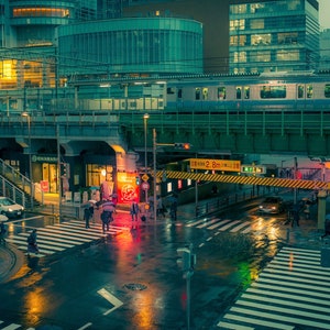 May include: A rainy night scene in a Japanese city with a train passing over a street intersection. The street is wet and reflective, with pedestrians crossing the street. The train is silver and green, and the buildings are a mix of modern and traditional styles.