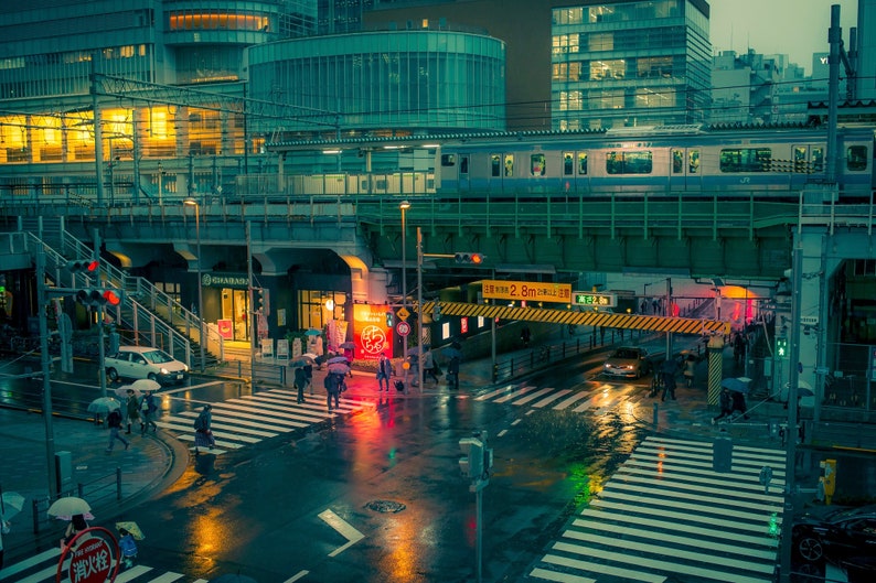 May include: A rainy night scene in a Japanese city with a train passing over a street intersection. The street is wet and reflective, with pedestrians crossing the street. The train is silver and green, and the buildings are a mix of modern and traditional styles.