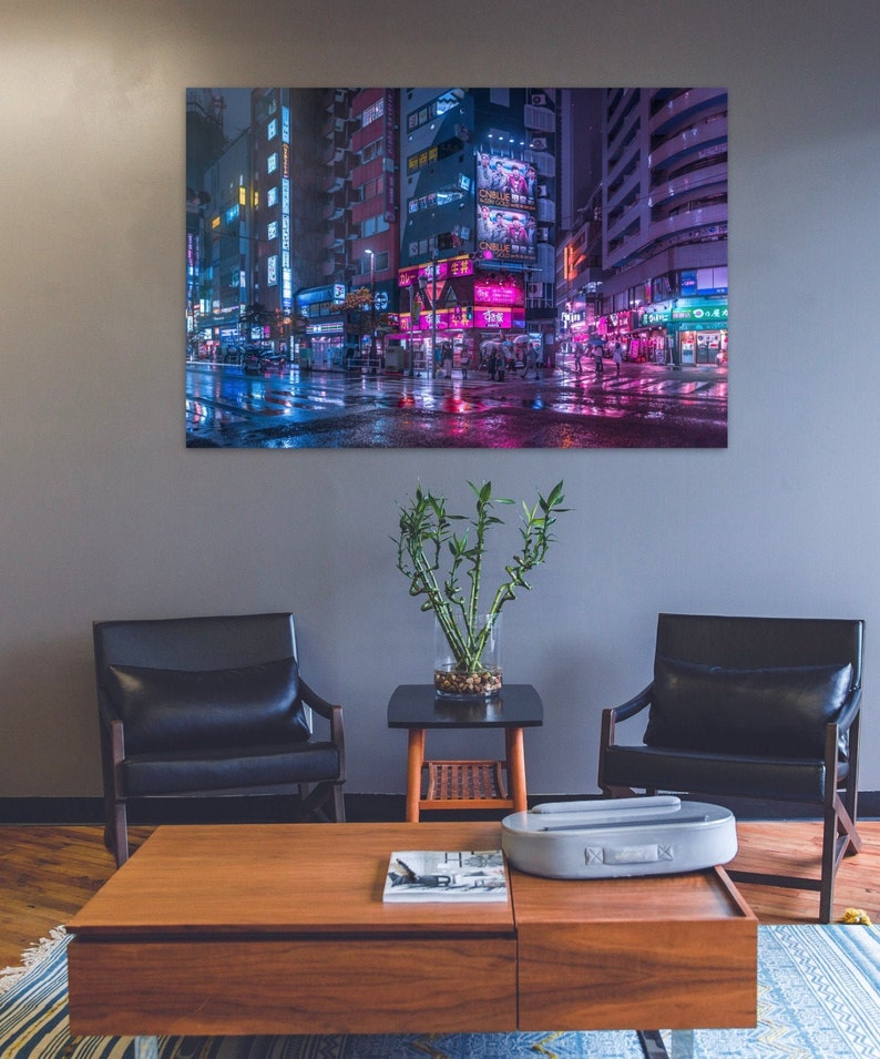 Pu&ograve; includere: Un interno di un soggiorno con due poltrone in pelle nera, un tavolino da caff&egrave; in legno, una pianta in vaso e una stampa incorniciata di una strada cittadina illuminata al neon di notte. La stampa presenta una strada bagnata con i riflessi delle luci al neon.