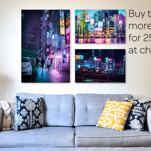 May include: Three framed prints of cityscapes at night with neon lights. The prints are hanging on a white wall above a gray couch with pillows. The text "Buy two or more prints for 25% off at checkout" is in the upper right corner of the image.