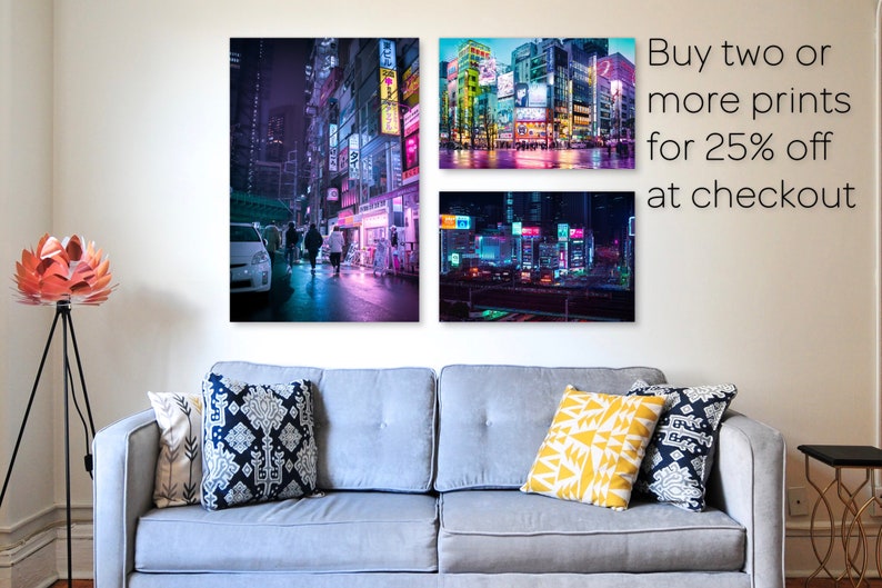May include: Three framed prints of cityscapes at night with neon lights. The prints are hanging on a white wall above a gray couch with pillows. The text "Buy two or more prints for 25% off at checkout" is in the upper right corner of the image.