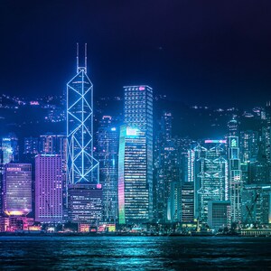 May include: A night view of a city skyline with tall buildings lit up in blue and purple lights. The buildings are clustered together and reflect in the water below.