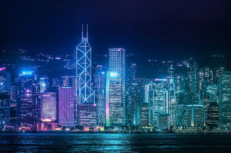 May include: A night view of a city skyline with tall buildings lit up in blue and purple lights. The buildings are clustered together and reflect in the water below.
