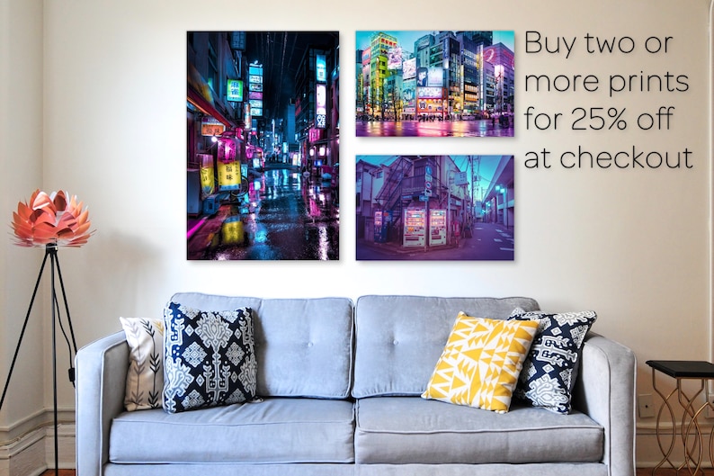 May include: Three framed prints of neon-lit cityscapes at night. The prints are hanging on a white wall above a gray couch with pillows. The text "Buy two or more prints for 25% off at checkout" is in the upper right corner of the image.