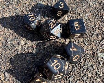 May include: A set of black dice with gold numbers. The dice are made of a polished stone material and have a variety of shapes, including a d4, d6, d8, d10, d12, and d20.
