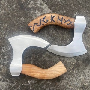 May include: Two axes with wooden handles. One axe has a silver blade and a wooden handle with a carved design. The other axe has a silver blade and a wooden handle with a carved design and the text "KNY" in black.
