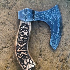 May include: A hand-crafted axe with a wooden handle carved with intricate designs. The axe head is decorated with a tree and a knotwork pattern.