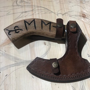 May include: A brown leather axe sheath with a decorative stitch pattern and a brass button. The sheath is attached to a wooden axe handle with the initials "MM" carved into the wood.