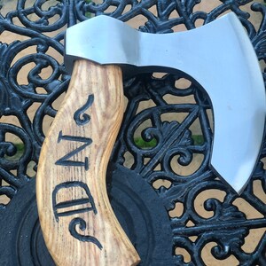 May include: A small, hand-forged axe with a wooden handle. The handle is carved with the words "N-D" and has a natural wood grain. The axe head is made of polished metal and has a curved blade.