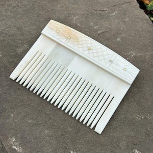 Puede incluir: Un peine de hueso blanco con un mango decorativo. El mango tiene un diseño tallado y detalles dorados.