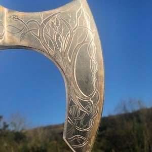 May include: Close-up of a silver axe head with an intricate design of a tree and vines etched into the surface.