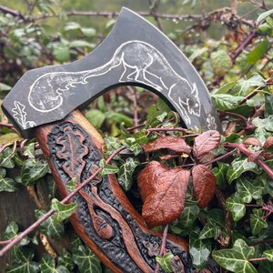 May include: A hand-carved wooden axe head with a black handle. The axe head is decorated with a carved image of an oak leaf and acorns. The handle is decorated with a carved image of a squirrel.