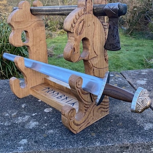 May include: A wooden sword and axe display stand with carved horse head details. The stand holds a silver and brown sword with a decorative hilt and a small axe. The word "TRYNDBIA" is carved into the base.