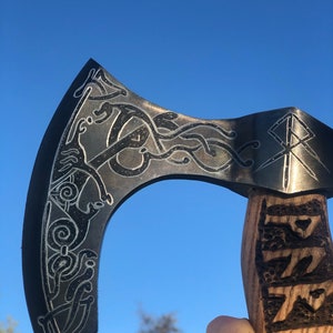 May include: A black axe with a wooden handle. The axe head is decorated with intricate carvings and the handle is carved with a pattern. The axe is held against a blue sky.