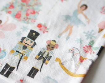May include: White fabric with a repeating pattern of Christmas-themed illustrations. The design includes ballerinas, nutcrackers, snowflakes, and floral elements in shades of blue, pink, and gold. The fabric has a soft, textured appearance.