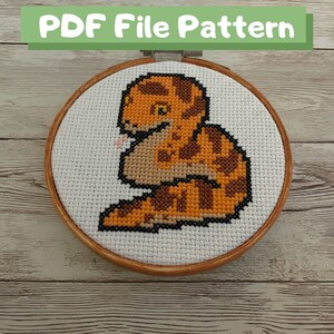 May include: A cross-stitch pattern of a brown and orange snake with a white background. The snake is curled up in a circle and has its tongue sticking out. The pattern is framed in a wooden hoop.
