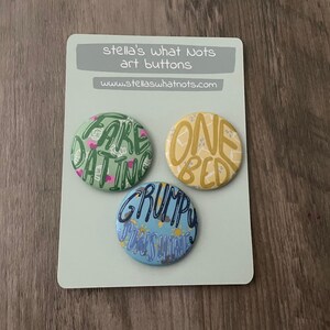 Könnte beinhalten: Drei runde Knöpfe mit bunten Designs und Text. Die Knöpfe zeigen "Fake Dating", "One Bed" und "Grumpy Was Right".