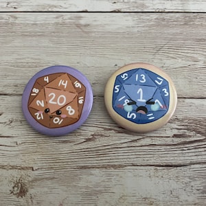 May include: Two round, colorful buttons with cartoon dice designs. One button is purple with a brown die showing the number 20. The other is blue with a die showing the number 1. Both dice have cute, crying faces.