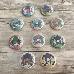 Op de afbeelding: Tien kleurrijke ronde pinback-knopjes met cartoon-spinnen met verschillende gekleurde lichamen en poten. De spinnen staan allemaal in verschillende poses, sommige kruipen, sommige springen en sommige staan stil. De knopen zijn allemaal 2,5 cm in diameter en perfect om een ​​tikje whimsy toe te voegen aan je rugzak, jas of draagtas.