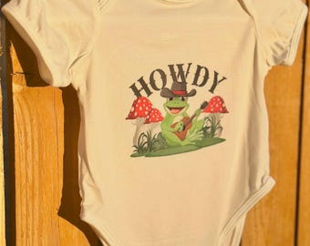 May include: Cream-colored baby onesie with a graphic of a green frog wearing a cowboy hat and playing a guitar. The word "HOWDY" is printed above the frog. Red and white mushrooms are on either side of the frog.