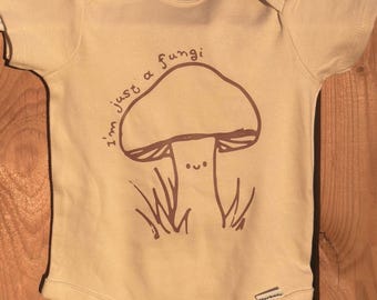 May include: A beige baby onesie with a brown mushroom graphic and the text "I'm just a fungi".