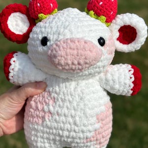 May include: A white crocheted stuffed cow with pink spots and red strawberry accents on its ears and head.