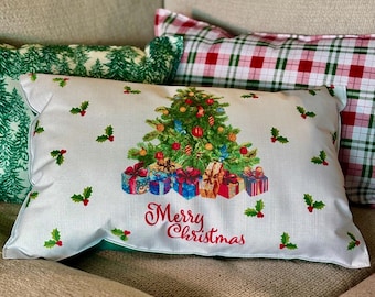 May include: A rectangular Christmas pillow with a decorated tree and wrapped gifts. The pillow has a white background with holly sprigs and the words "Merry Christmas" in red script. Other pillows are visible in the background.
