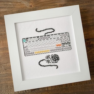 May include: A white framed cross-stitch with a black and white image of a cassette tape and a rose. The cassette tape has a gray and white pattern and the text "Cassette Tape" is stitched in black.