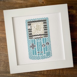 May include: A framed cross-stitch design of a teal blue handheld video game console with a black and white pixelated screen. The console has a directional pad, four buttons, and a start button.
