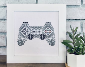 May include: A framed cross-stitch artwork featuring a detailed video game controller design. The controller is primarily gray and black, with accents of pink and blue. The artwork is displayed in a white frame, set against a brick wall.