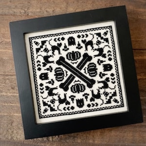 May include: Black and white cross-stitch embroidery framed in a black frame. The design features a repeating pattern of bones, cats, pumpkins, and bats.