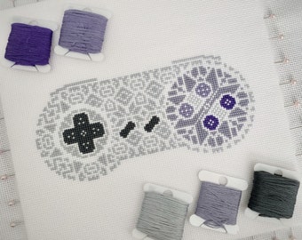 May include: A cross-stitch pattern of a video game controller with purple, gray, and black embroidery floss. The controller is on a white Aida cloth with pins holding the fabric in place.