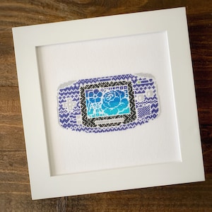May include: A purple and white cross-stitch of a handheld video game console with a blue and white pixelated image of a rose on the screen.