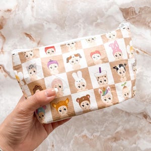 May include: A rectangular cosmetic bag with a zipper closure. The bag features a repeating pattern of cartoon-like faces with various animal and accessory designs on a brown and white checkered background.