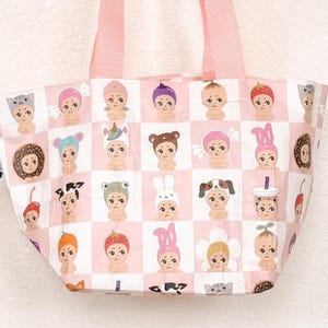 May include: Pink tote bag with a repeating pattern of cartoon baby faces wearing various hats and accessories. The bag has pink handles and a light pink and white checkered background. The bag is held by a hand.