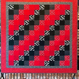 Può includere: Una trapunta rossa, nera e bianca con un motivo geometrico. La trapunta è appesa a un muro di mattoni.