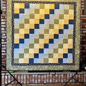 Può includere: Una trapunta fatta a mano con un motivo a scacchiera nei toni del giallo, blu e verde. La trapunta ha un bordo floreale ed è esposta su un supporto in metallo nero. È un accogliente oggetto di arredamento per la casa.