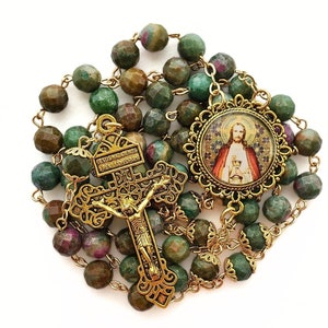May include: A green and gold rosary with a crucifix and a round pendant featuring an image of Jesus holding a chalice. The beads are made of green aventurine.