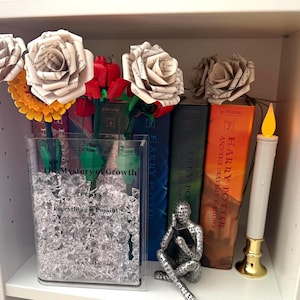 May include: A shelf display featuring paper flowers, books, and decorative items. A clear vase holds paper flowers and artificial ice. Books include "Harry Potter" titles. A candle and a metal figure complete the arrangement. The vase has the text "The Mystery of Growth" and "Everything is Possible".